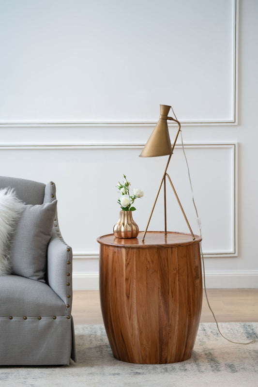 A wooden drum side table with a tray-style top and a small vase with flowers on top, positioned next to a sofa in a minimalistic setting.