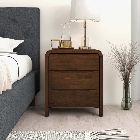 Solid wood nightstand with three drawers, placed next to a bed with grey headboard in a neutral room setting.
