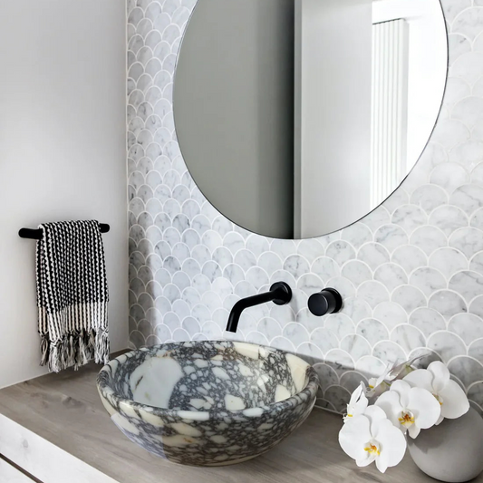 A Calacatta Viola marble vessel sink above a vanity with a black faucet and a fishscale mosaic tile wall behind it, set in a modern bathroom with a black and white hand towel and white orchids on the counter.