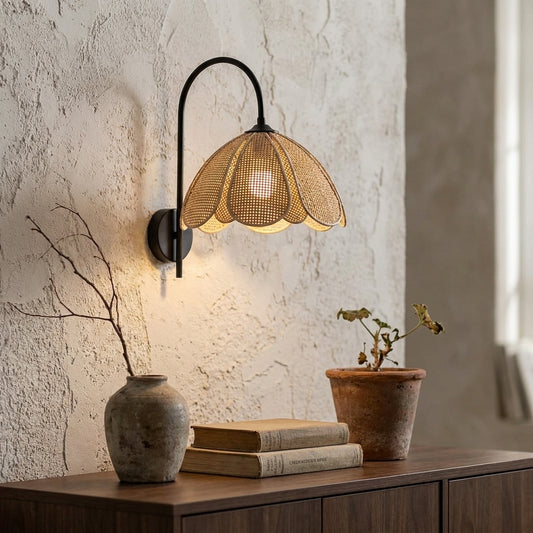 A handwoven rattan flower-shaped wall sconce with a black metal arm installed on a textured wall above a wooden surface with books, a vase, and a potted plant.