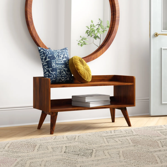 A wooden storage bench with open shelves, holding books, pillows, and a mirror in the background, under a decorative round mirror on white walls and patterned rug