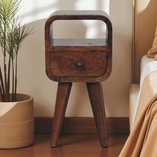Dark wood extra mini curve bedside table with a drawer, placed on a wooden floor near a potted plant and a bed with golden sheets.