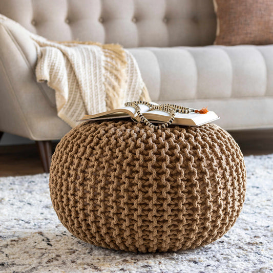 A beige knitted pouf with a rope-like texture, atop a white rug in a living room setting with furniture in the background.