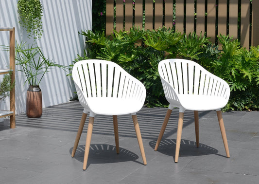 Two white Nassau Chairs with resin and wood finish on a paved patio with plants and a fence in the background