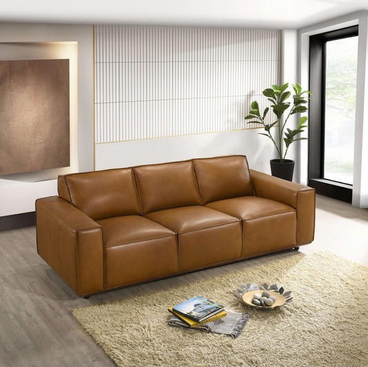A tan leather sofa with wooden legs, placed on a shaggy rug in a minimalist room with a potted plant nearby and a large window in the background.