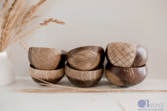 Six coconut bowls arranged with decorative patterns, placed against a white vase with wheat stems, displayed on a table with a neutral background.