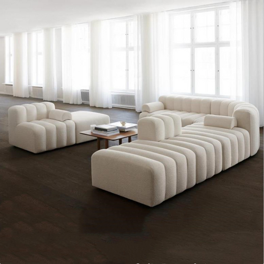 Sculptural curved sofa in cream fabric with modern design, light wood coffee table and large windows in background.