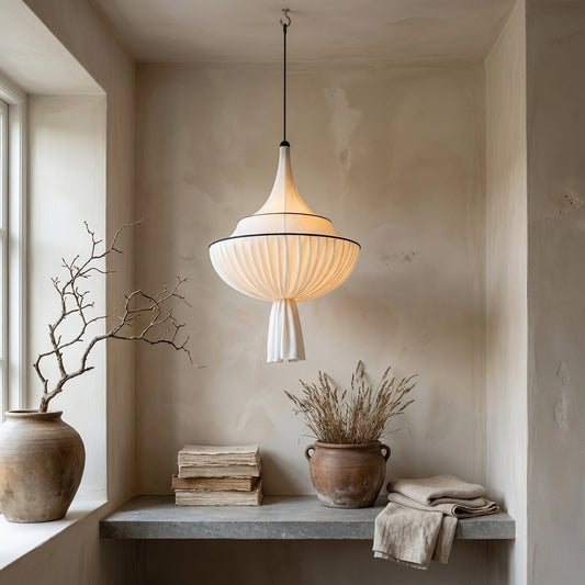 Bell-shaped pleated fabric pendant light with a soft tassel hanging in a corner with a vase, stack of books, and potted plant below it.