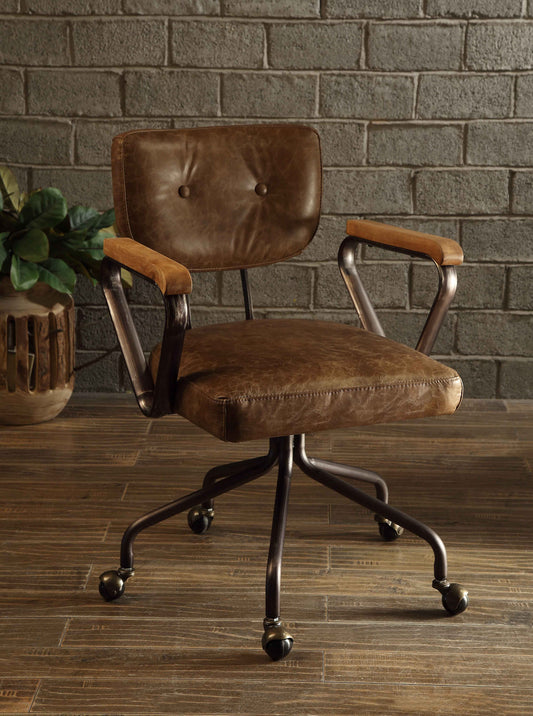Vintage-style office chair with leather upholstery, metal frame, and wooden armrests, positioned in front of a brick wall with a wooden floor and a plant in a rustic setting.