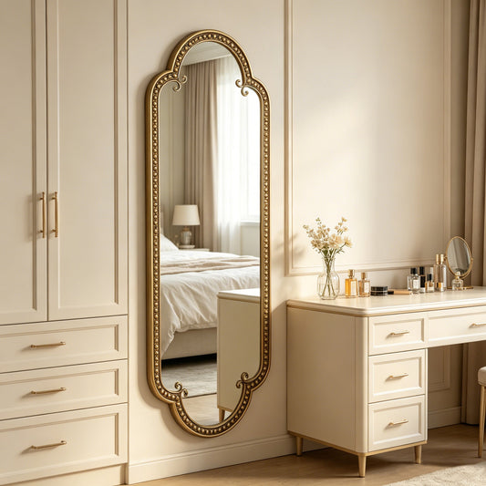 Elegant scalloped oval wall mirror with decorative gold frame, hanging against a cream-colored wall in a bedroom setting with a bed and dresser visible.