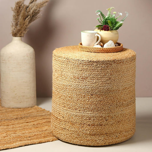 A round jute pouffe ottoman stool with a beige finish, atop a beige surface next to a decorative glass vase, showing an indoor setting.