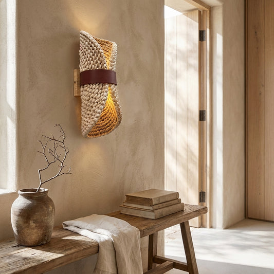 A handcrafted water hyacinth wall sconce with a rustic woven texture, light brown and natural tan tones, mounted on a beige textured wall above a wooden bench with books and a vase.