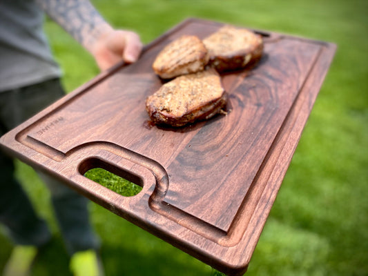 A walnut cutting board with handles and juice groove, being held outdoors with cooked pastries on it.