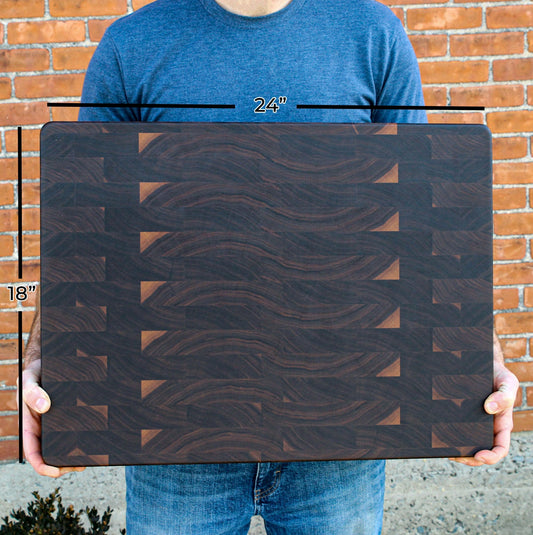 Man holding a large rectangular wooden cutting board with end grain texture, 24 inches by 18 inches, resting on his legs against a brick wall.