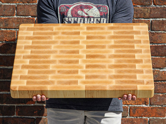 A person holding a XXL maple end grain cutting board with visible grain patterns and edges against a brick background, USA-made.
