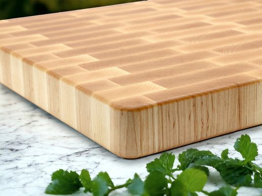 A natural maple cutting board with a distinctive diamond pattern and rubber feet, sitting on a white marble surface with herbs in the foreground.