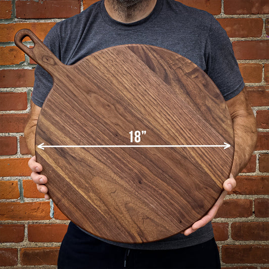 Person holding round walnut paddle-shaped cutting board with a handle, marked as 18 inches long, on a brick wall backdrop.