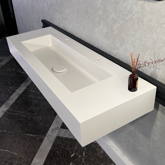 White quartz monolithic trough sink handcrafted in a modern setting with a dark marble floor and textured wall, containing a plug and a black vase with sticks and greenery.
