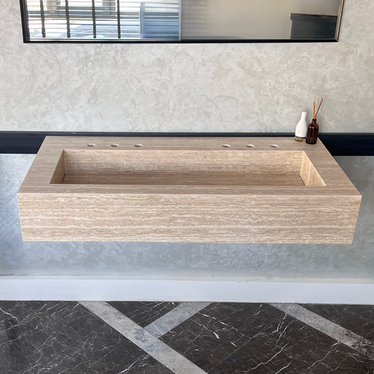 Rectangular handcrafted bathroom sink made of natural travertine with a rustic finish, placed on a floating mount against a textured wall, next to small bath accessories in a chic setting