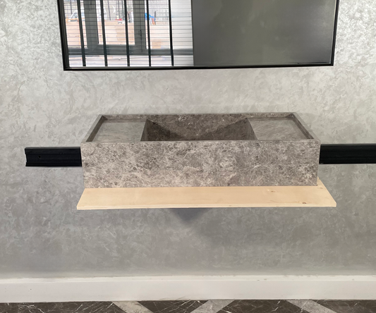 A rectangular tundra grey marble bathroom sink with a natural pattern, mounted on a light wood board against a textured concrete wall with a window above it.