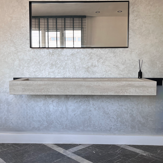A rectangular, beige porcelain sink mounted on a textured wall, with a hanging mirror above and a black vase containing sticks to the right of the sink in a modern, minimalist-style room with black and white marble floor tiles.