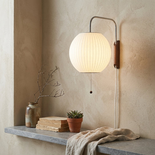 A minimalist wall sconce with a round gourd-shaped shade and pull chain, mounted on a textured beige wall. Below it, a shelf displays a vase, books, a terracotta pot with a succulent, and a draped cloth.