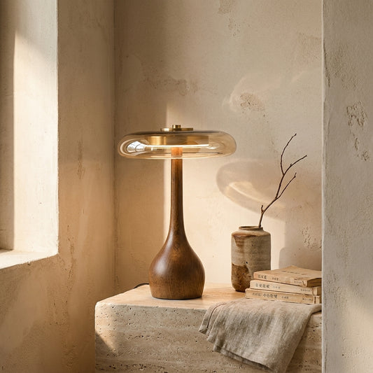 Modern wooden and glass table lamp in a minimalist setting against a textured wall, with sunlight streaming in through a window