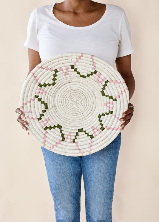 A person holding a woven wall basket with a sunburst pattern in white, pink, and green.