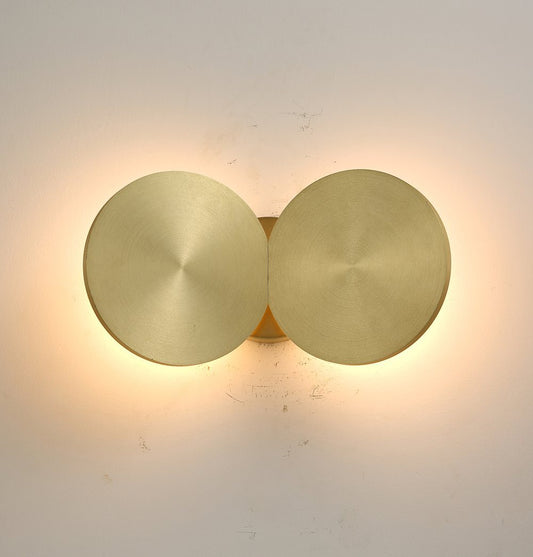 Two circular wall lights with a brushed brass finish, emitting a warm glow against a white wall.