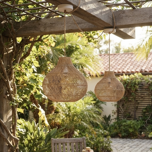 Two rattan pendant lights hanging on a pergola in an outdoor setting with greenery and a tiled roof in the background.
