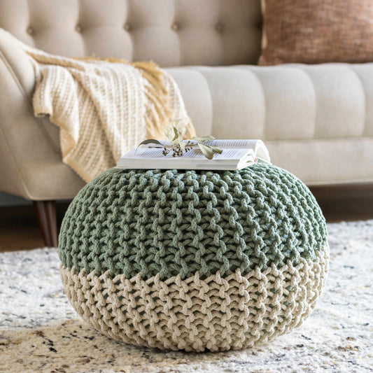 Circular knitted pouf, grey and beige, on a patterned rug in front of a sofa with a blanket and pillow.