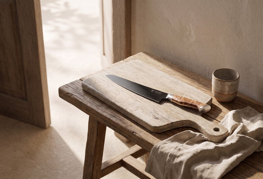 10" slicing knife on a wooden chopping board with a wooden handle, with a small ceramic cup and linen cloth nearby in a rustic setting