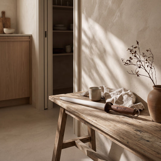 A wooden bench with a knife, cup, and tea towel; natural light with shadows on wall; rustic tones.