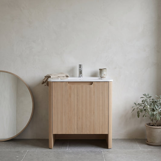 24 inch wall-mounted floating bathroom vanity made of light wood with vertical slats and a white countertop, against a neutral wall with a round mirror and a potted plant, no text overlay.