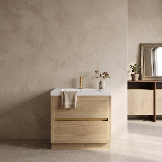 Wall-mounted bathroom vanity with two drawers, light wood finish, white sink, brass faucet, beige towel, neutral wall, concrete floor, potted plant visible on adjoining surface.