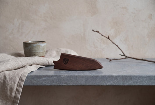 {"alt":"Wooden knife sheath; dark walnut finish, sits on a gray concrete surface with a beige cloth and a green ceramic cup in the background; no text overlay o