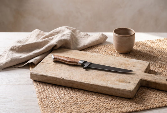 6" Boning Knife on a rugged wooden chopping board with a linen cloth and a ceramic mug in a minimalist setting.