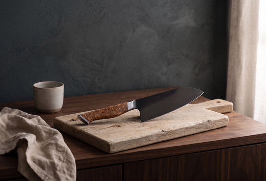 6 inch chef knife with wooden handle, on rustic wooden cutting board, beside ceramic cup, beige fabric and curtains, dark textured wall in background.