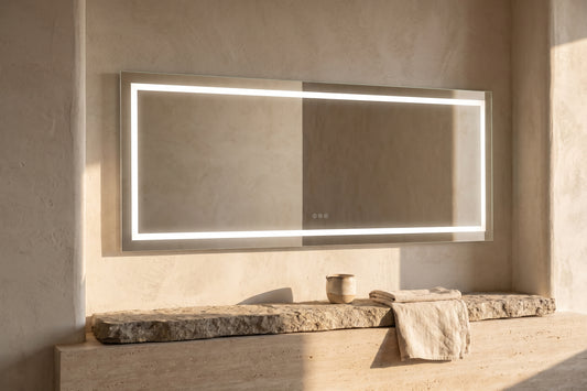 A large, sleek LED bathroom mirror with front and backlighting mounted in a minimalist bathroom setting. The mirror frame is metallic and the glass is clear. The surrounding walls are textured and neutral in color. Below the mirror is a stone counter with towels and a vase.