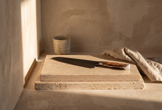 Wooden cutting board with a chef's knife resting on it, positioned on a stone slab with soft lighting surrounding the scene.