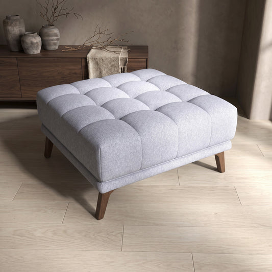 An Addison Square Upholstered Ottoman with tufted pattern and wooden legs, placed on wood flooring near a wooden console table.
