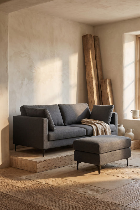 Dark grey sofa set with ottoman on wooden platform in minimalist room with natural light from window, wood panels, terracotta decorations.