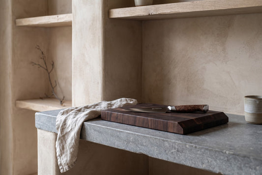 Wooden cutting board and knife on concrete countertop with textured beige wall and shelves in background