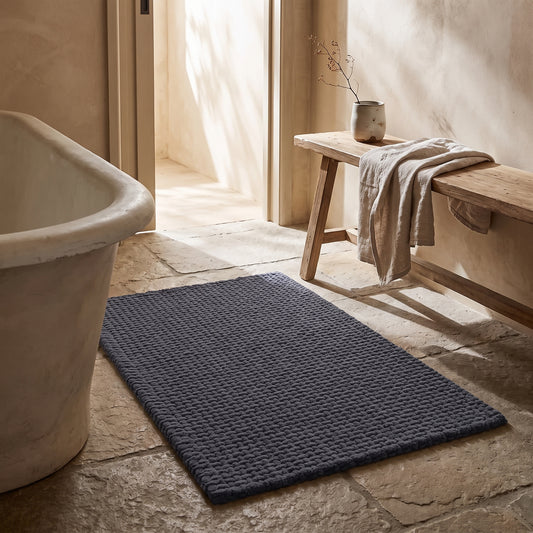 Dark gray chenille bath mat with textured surface on stone floor near bathtub.
