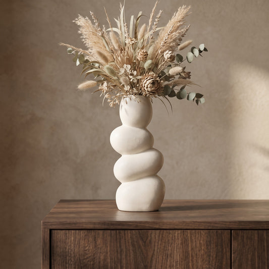 White pottery vase with a textured finish, topped with dried pampas grass and greenery, placed on a wooden table against a neutral wall.