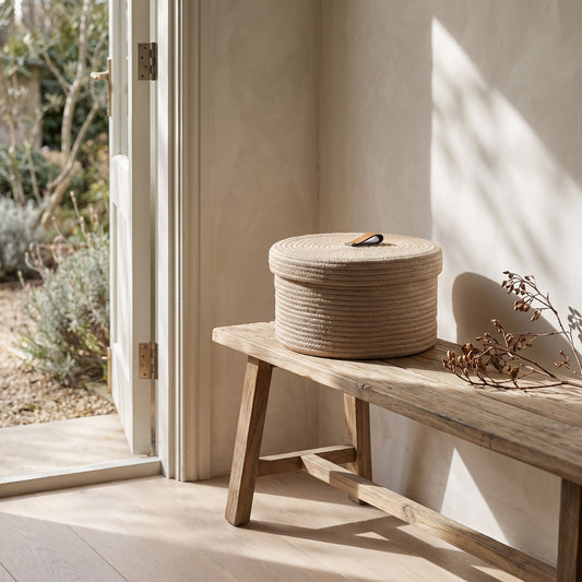 Circular jute basket with a lid next to a wooden bench in a light-filled room with a door to a garden outside.