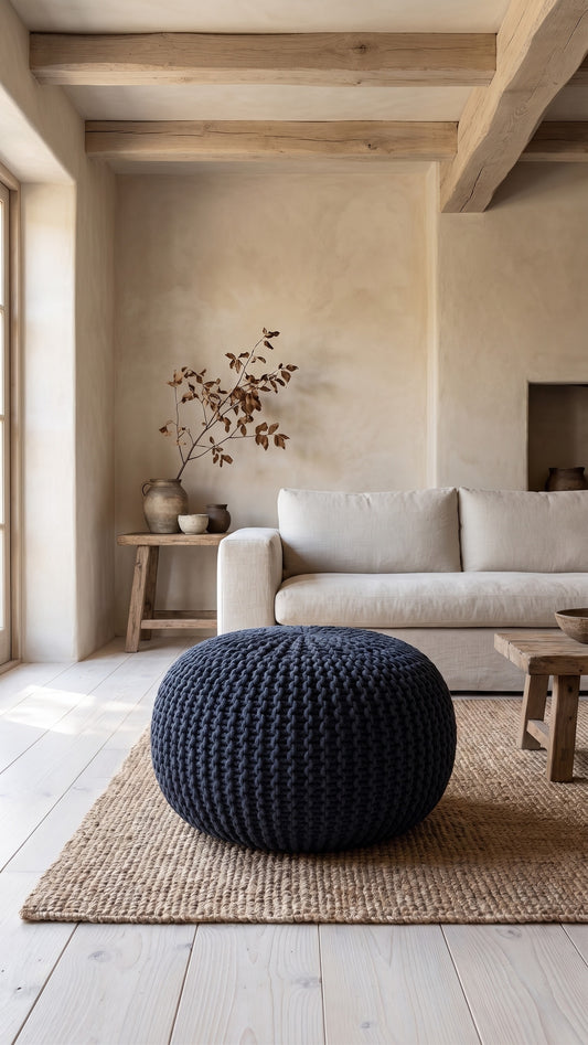 Dark blue knitted cotton round pouf ottoman on a woven rug in a minimalist living space with wood beams and natural light.