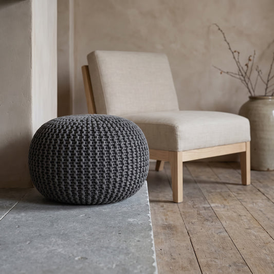A textured gray pouf in the foreground with a beige chair and vase in the background on a wooden floor.