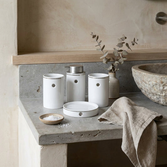 A four-piece bathroom set with white clay containers on a gray concrete counter, a bowl of salt, a folded towel, and a plant vase in the background.