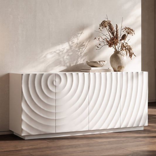 White buffet cabinet with textured panel design on a wooden floor, simple vase of dried flowers, books, and decorative wall shadow behind